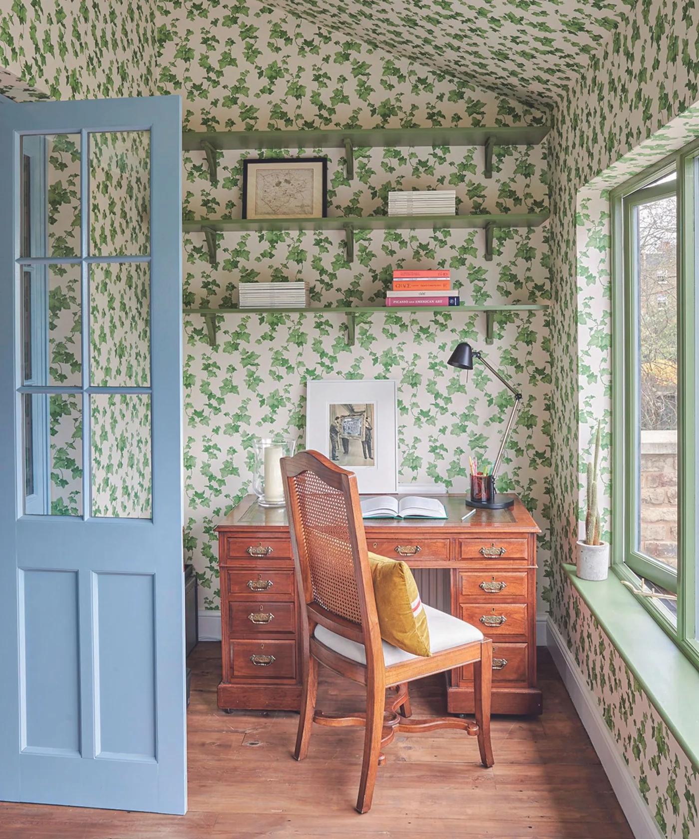 Small office covered in green ivy wallpaper with a wooden drawer desk and warm metal handles.