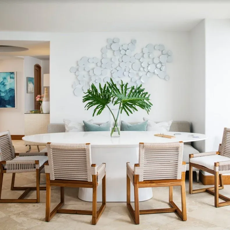 Clear vase with broad split green leaves on a white oval table surrounded by woven-back wood chairs