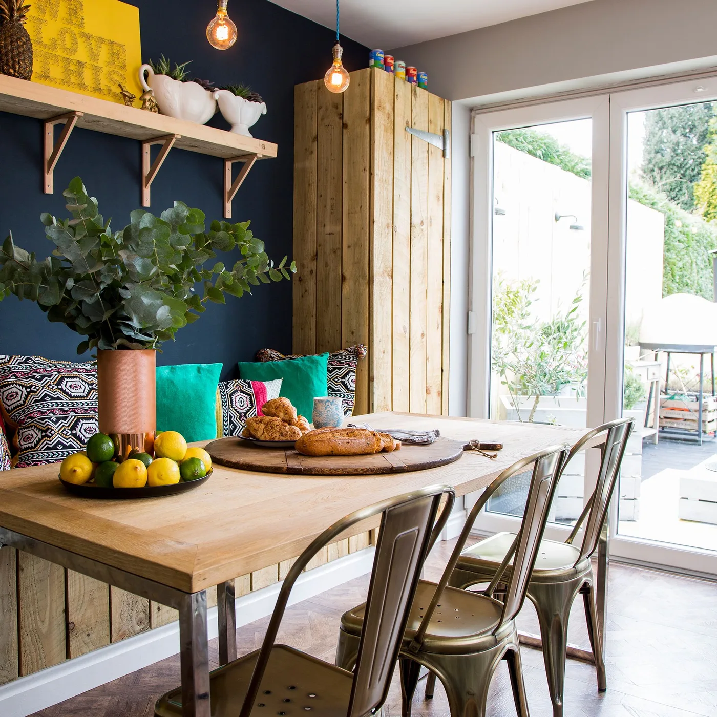 Country dining nook with wooden table, cushioned bench against a dark blue wall, metal chairs and glazed doors opening toward the garden.
