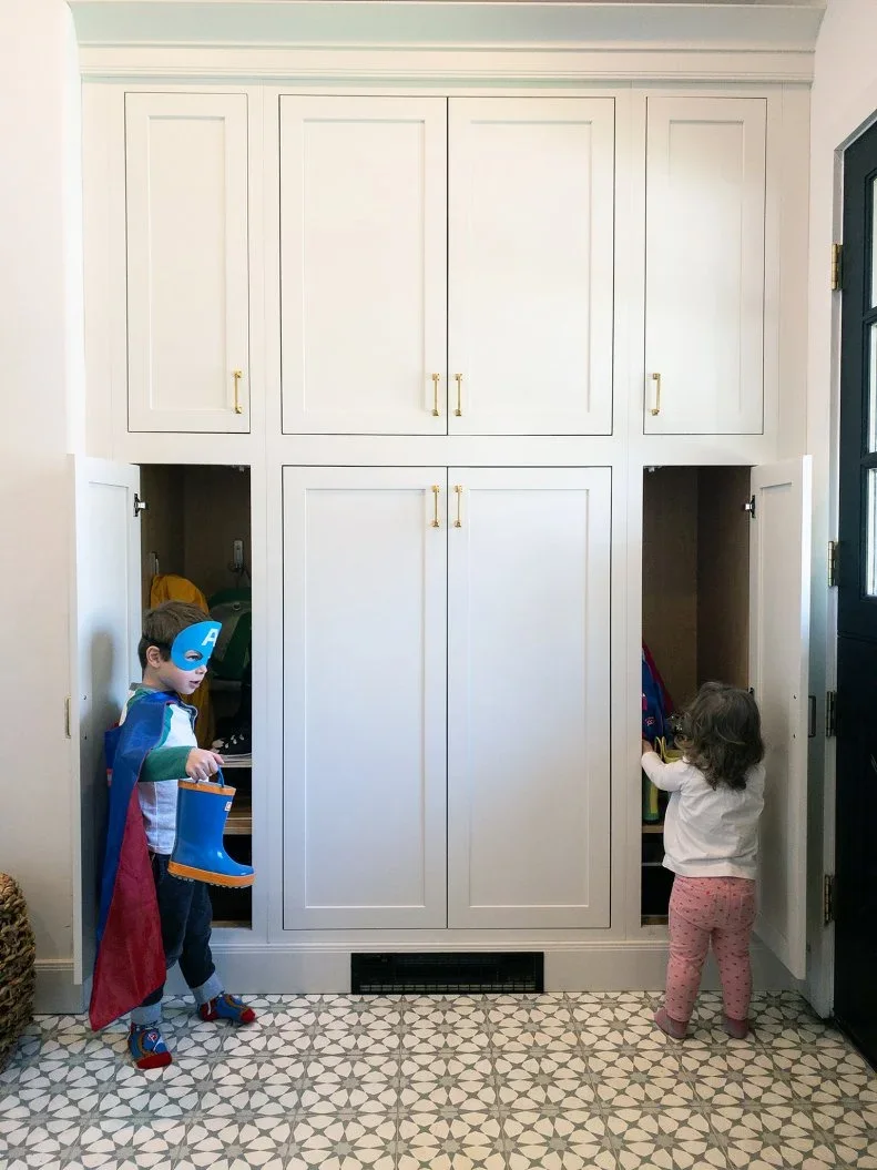 Tall pale framed entry cupboards with warm-toned handles stand beside dark door trim above a small repeating floor pattern, with lower cabinet doors open
