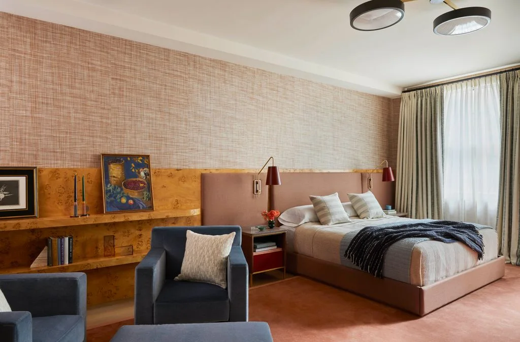 A dusty pink bed stands beside integrated wood shelves and red wall lamps beneath a textured pale wall, with dark chairs and light curtains nearby