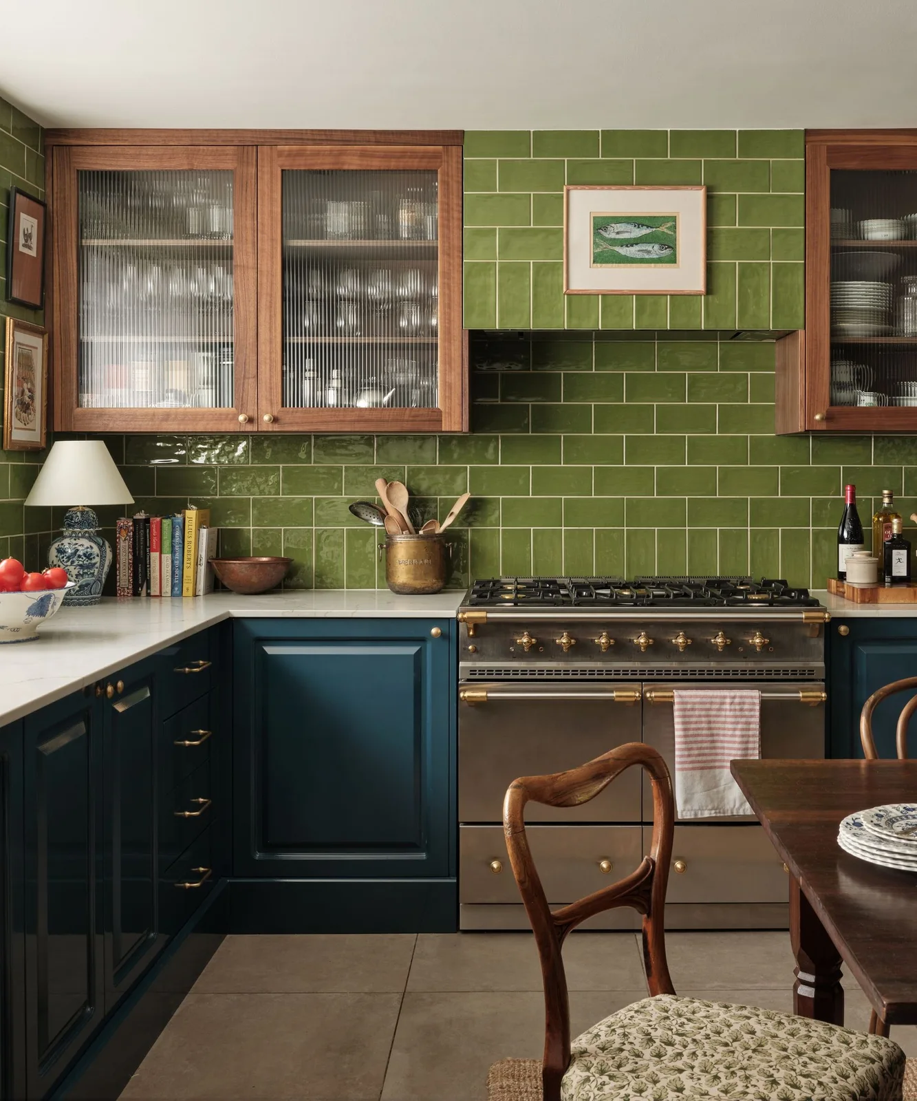 Olive green rectangular tiles surround wooden glass-fronted cupboards and a metal range above blue lower cabinet fronts