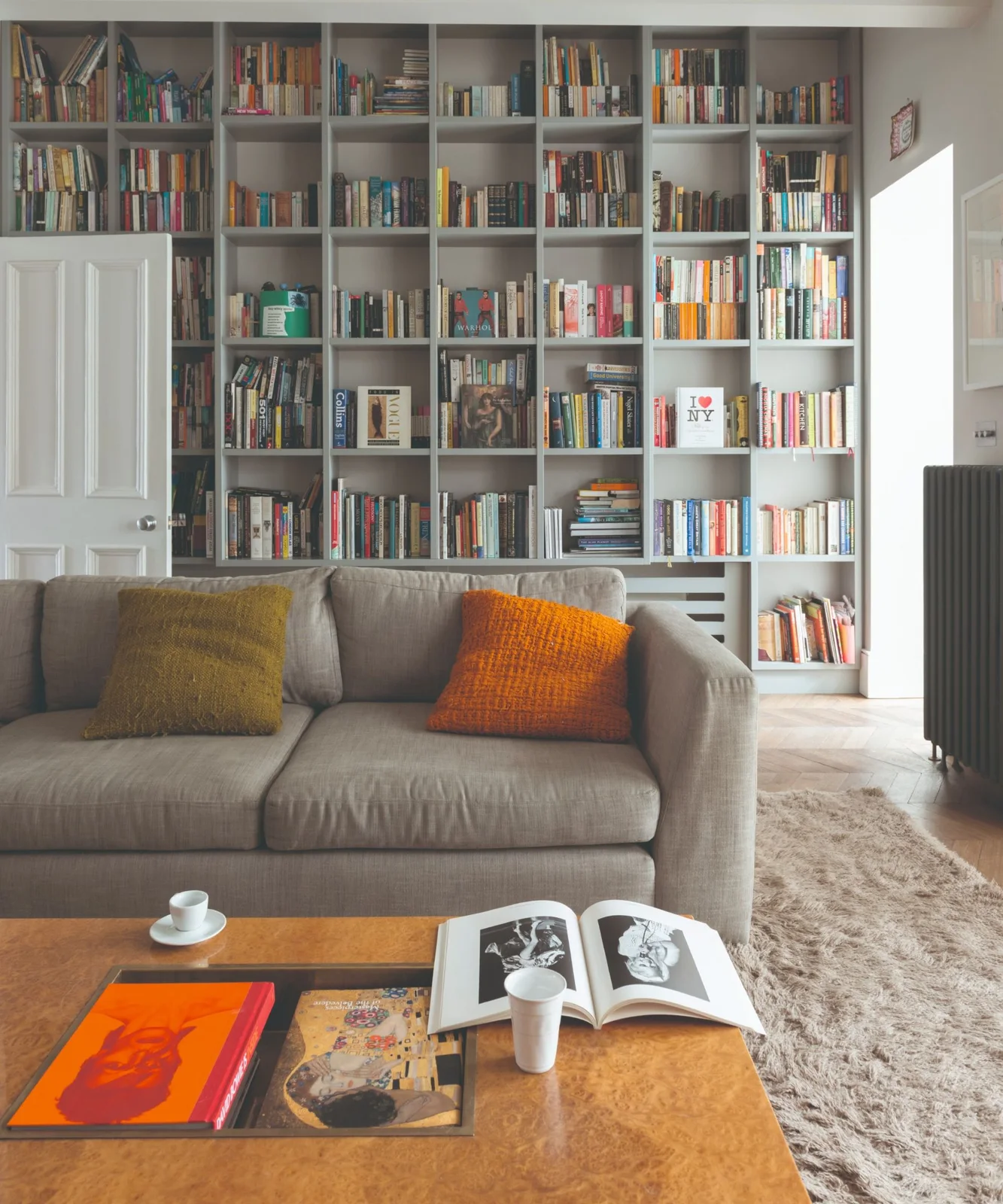 A gray sofa with mustard and orange cushions sits before tall white bookshelves beside a low wooden table and a textured light rug
