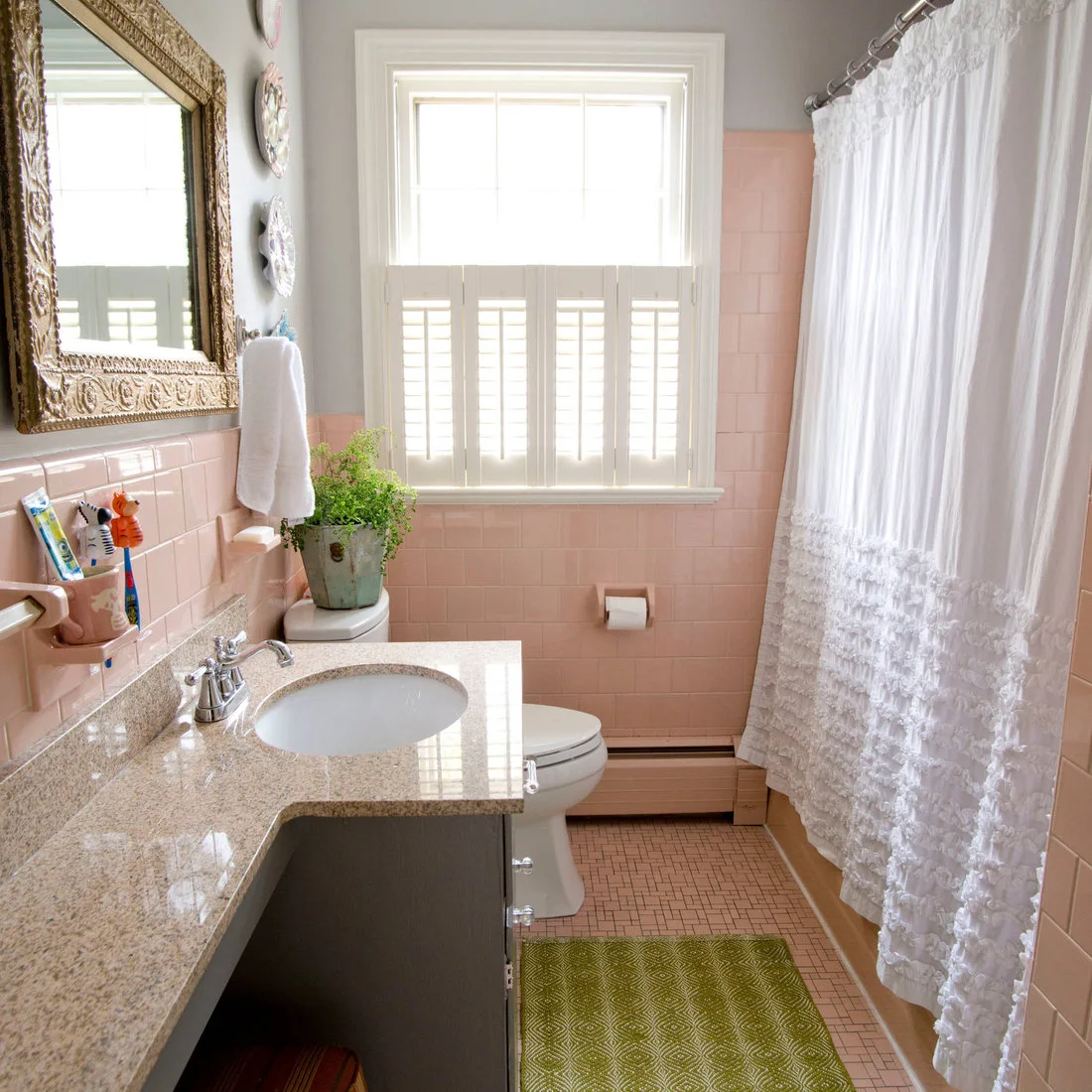 Pink wall tiles surround a bath and sink beside a white shower curtain, pale vanity counter and gold-framed mirror