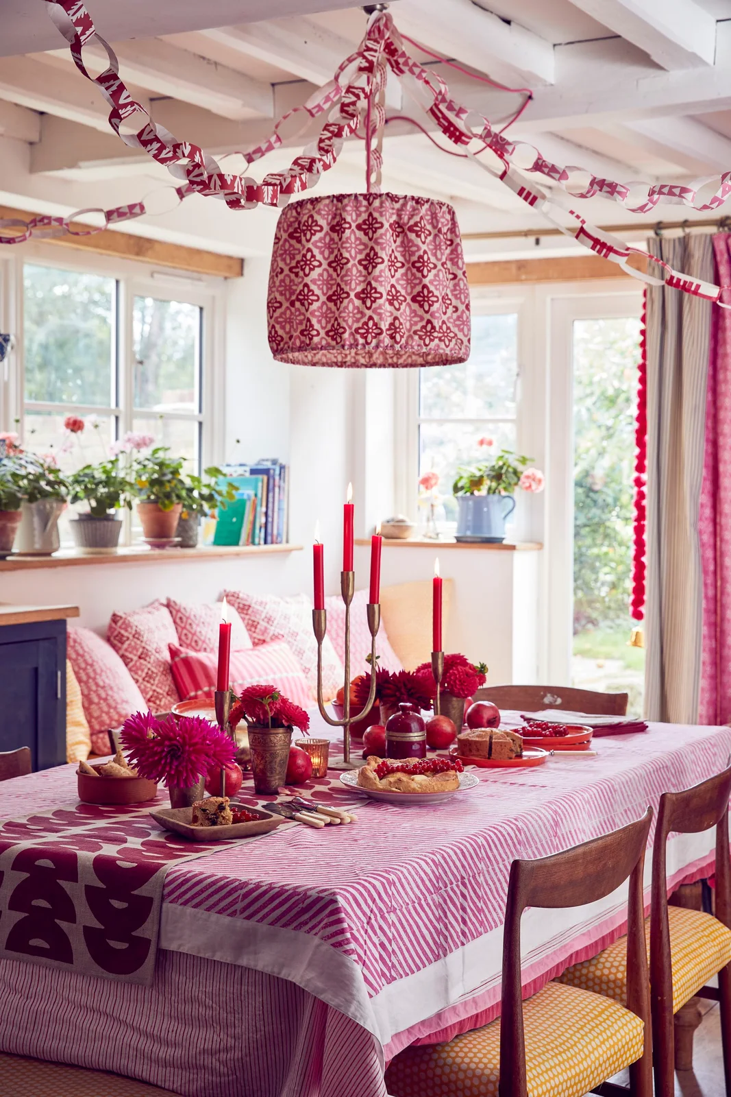 Pink patterned table linens sit beneath a patterned shade and paper chains beside yellow checked chair seats and red decorative accents
