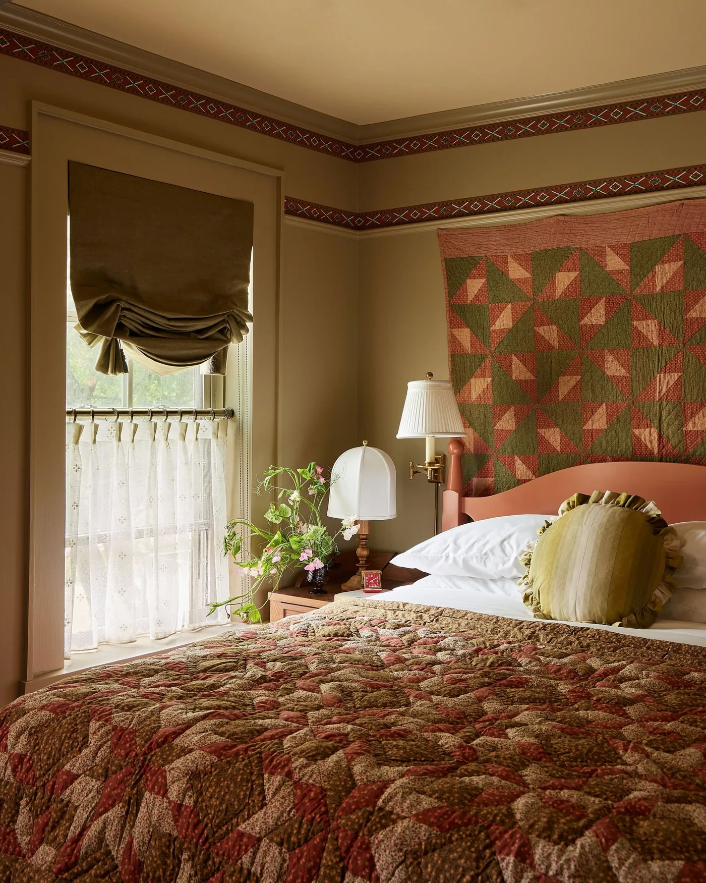 Warm bedroom with patchwork bedding, a geometric quilt hanging above the bed, patterned wall borders and shaded bedside lamps.
