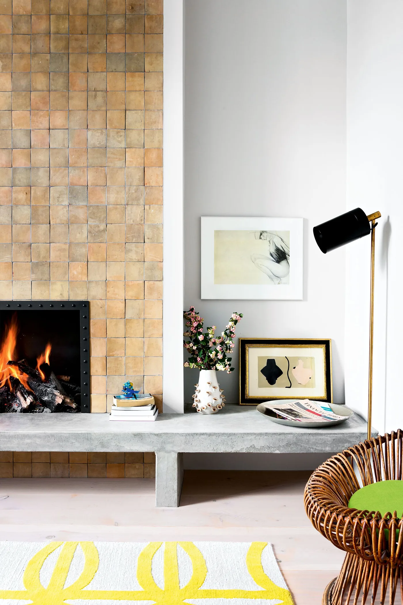 Small warm square tiles cover a tall fireplace wall beside a projecting pale hearth, framed pictures and a woven chair