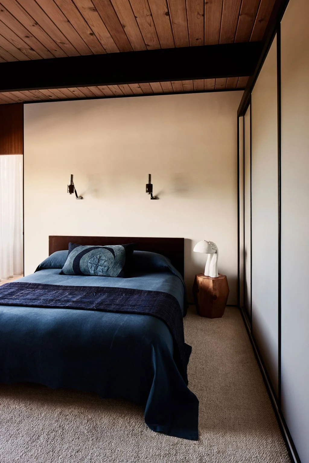 A dark blue bed stands below a wood-board ceiling with dark beams beside plain pale walls, small wall lights and a wood side table