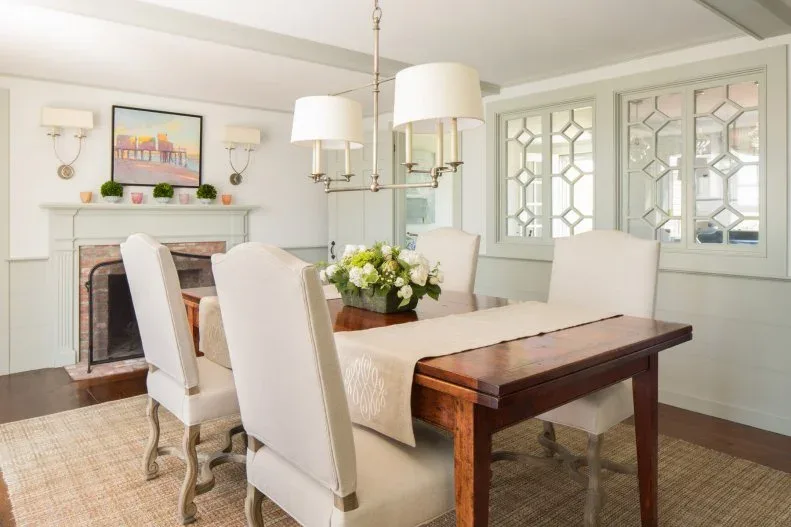 Dining room with a pale side wall containing three geometric framed mirrors, wooden table, cream chairs, fireplace, pale shaded lighting, and woven rug.