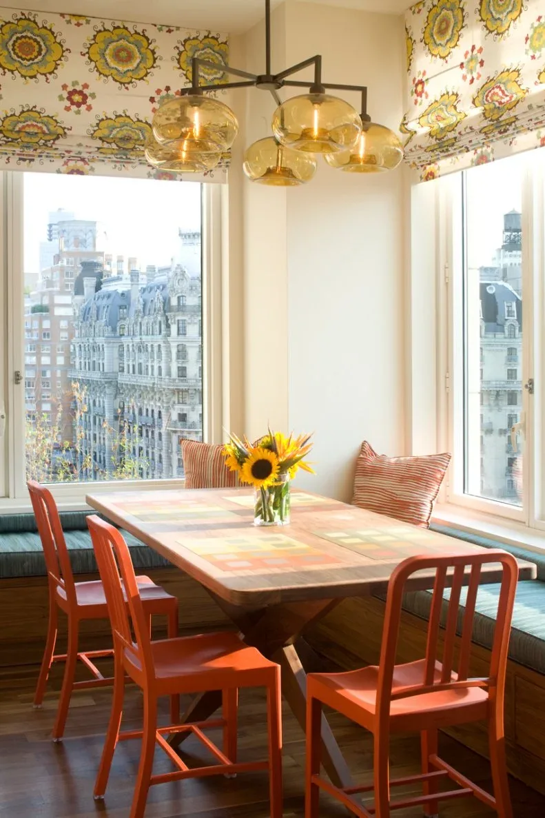 Corner window banquette with teal seat cushions, orange chairs, a wooden rectangular table, floral window shades, striped pillows, and amber-colored glass pendant shades.