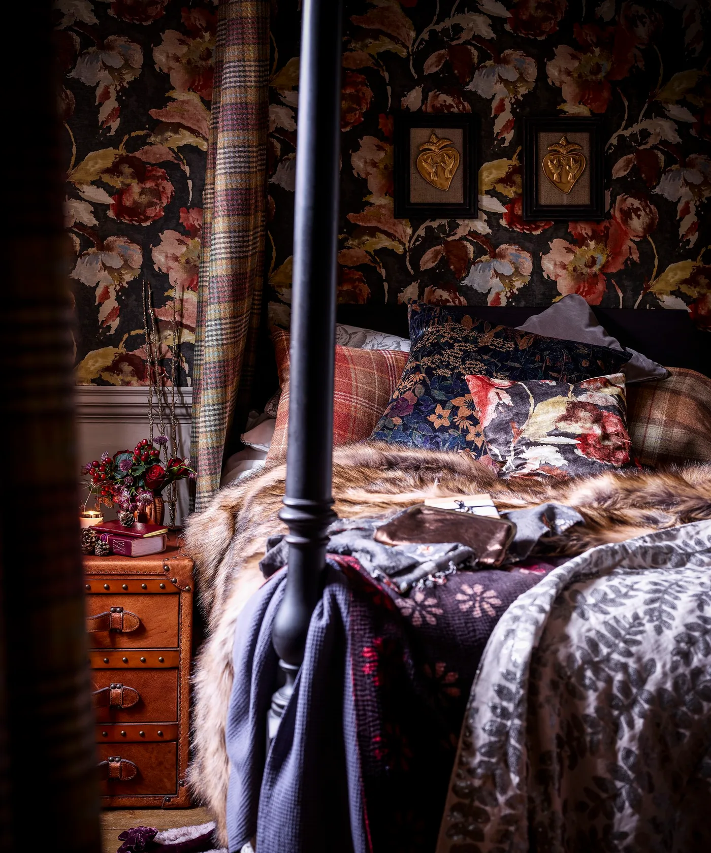 A dark four-poster bed sits against floral wallpaper with layered patterned bedding, plaid curtains and stacked bedside trunks