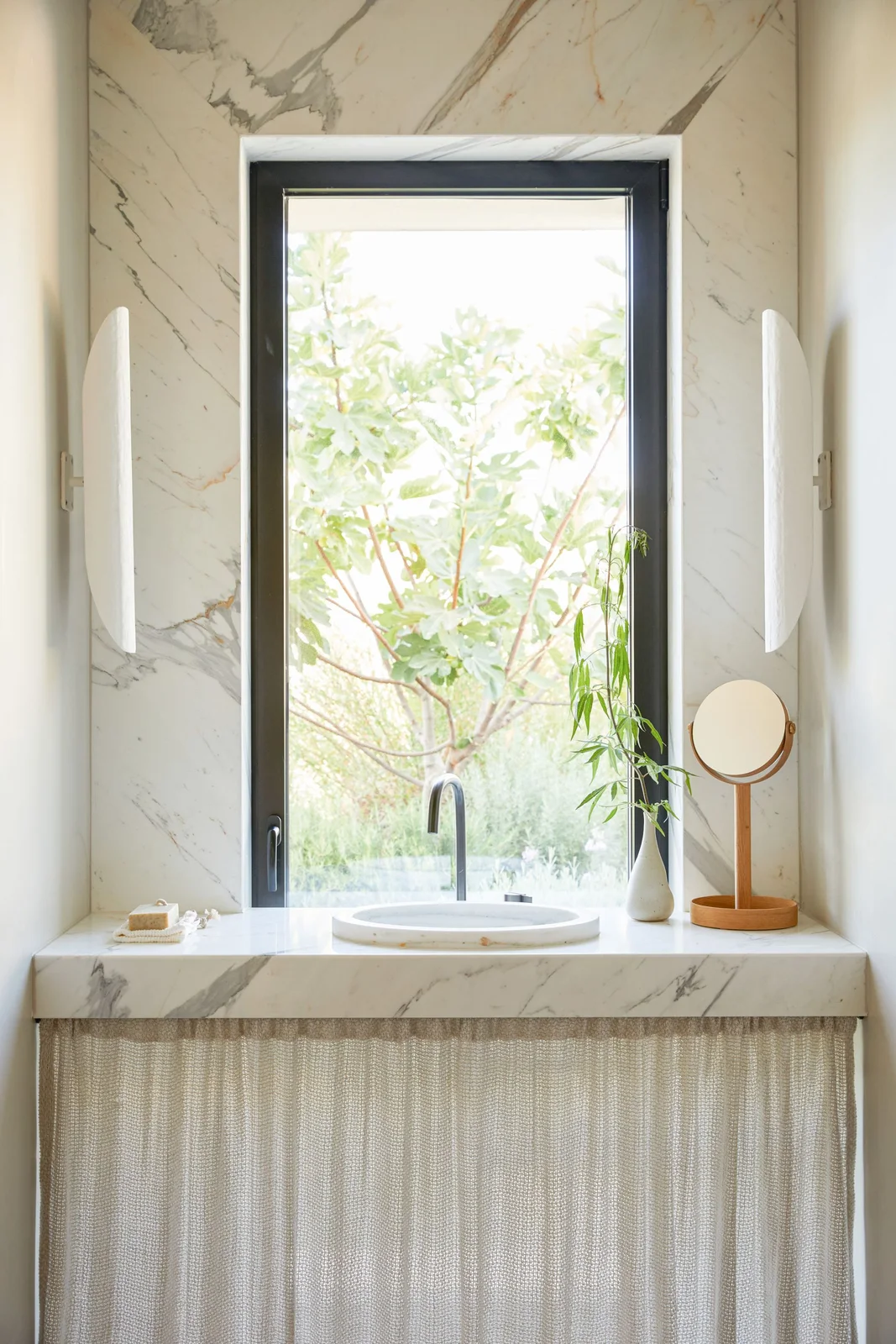 A pale veined counter with an inset round basin sits below a tall dark-framed window between slim pale wall lights, with a light curtain beneath