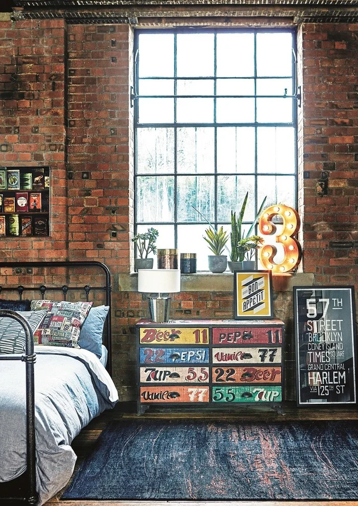 A dark metal bed sits beside a colorful drawer chest and large grid window against exposed brick with framed signs and small plants