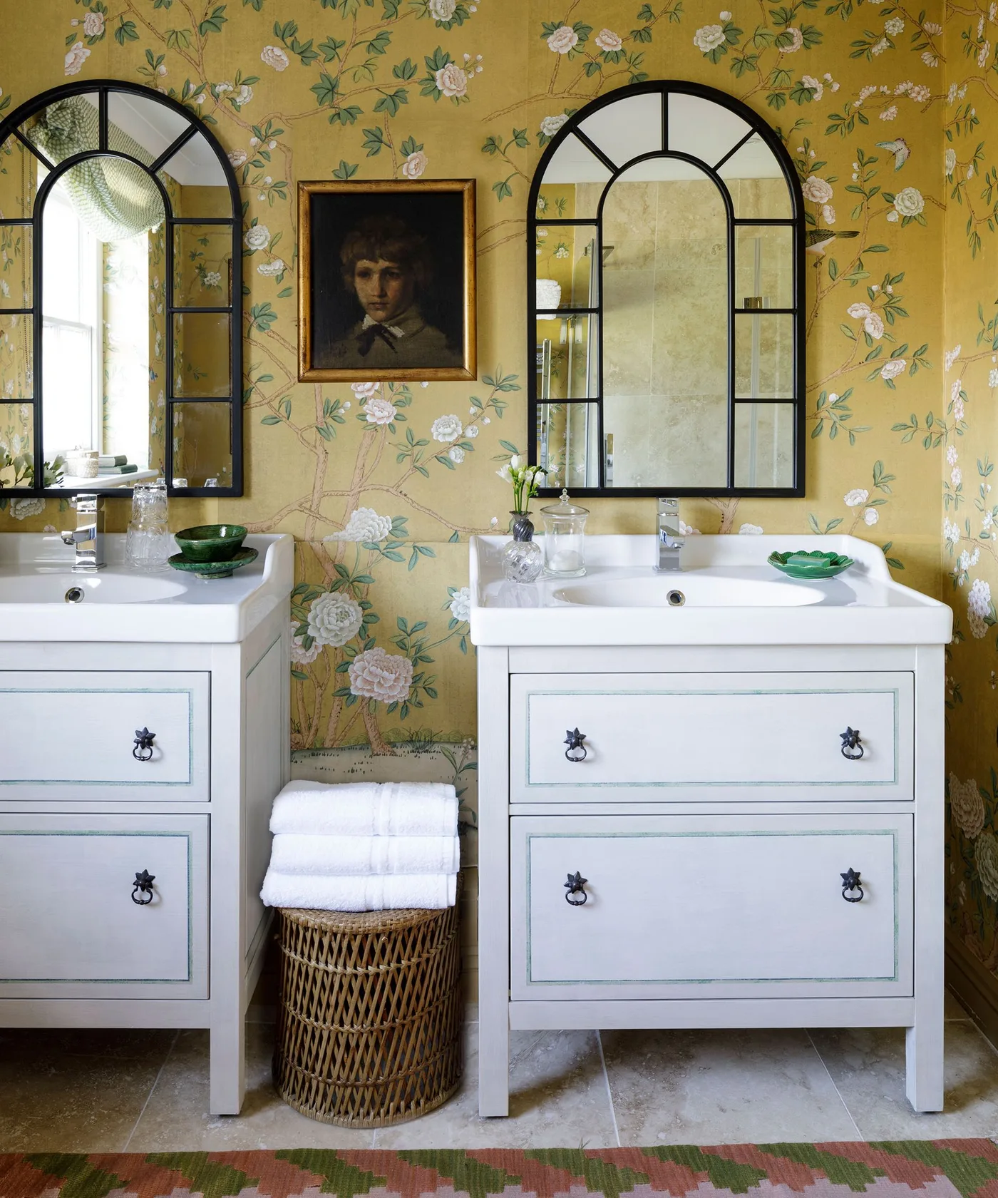 Country bathroom with yellow flowering-branch wallpaper, two pale vanities and dark arched mirrors