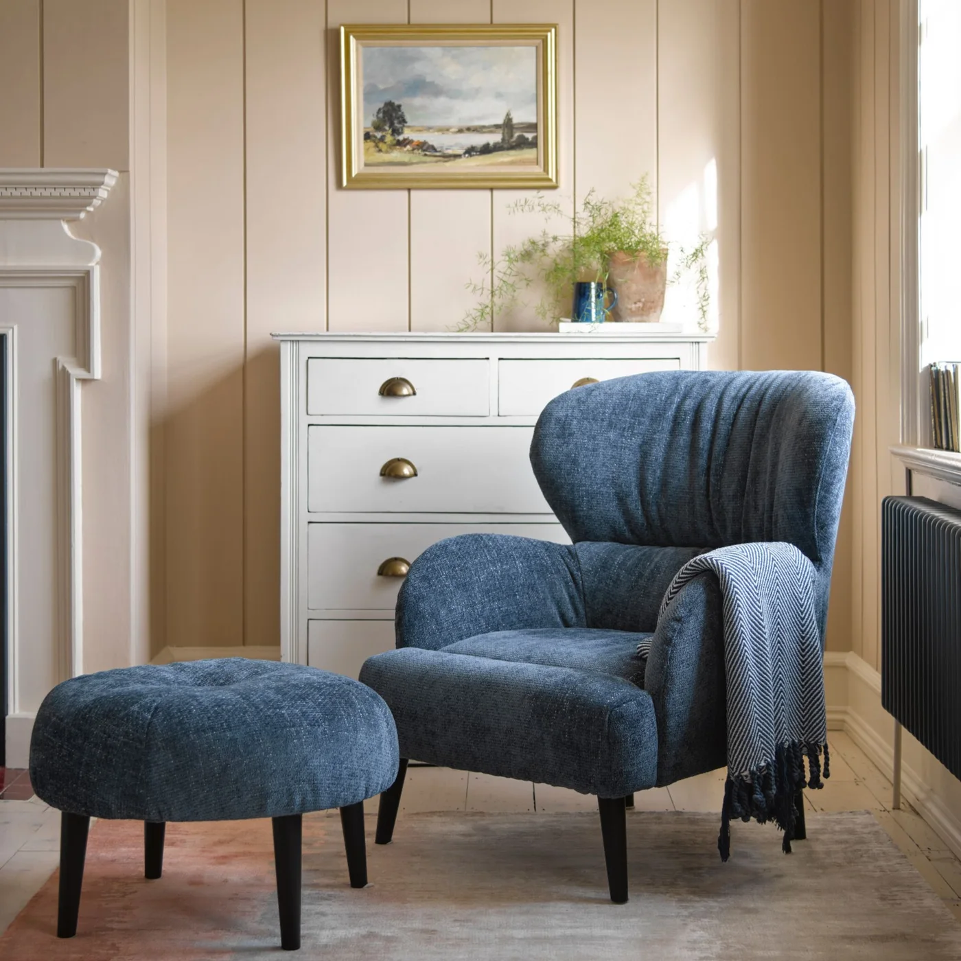 A blue rounded armchair and matching footstool sit beside a pale drawer chest under a framed landscape