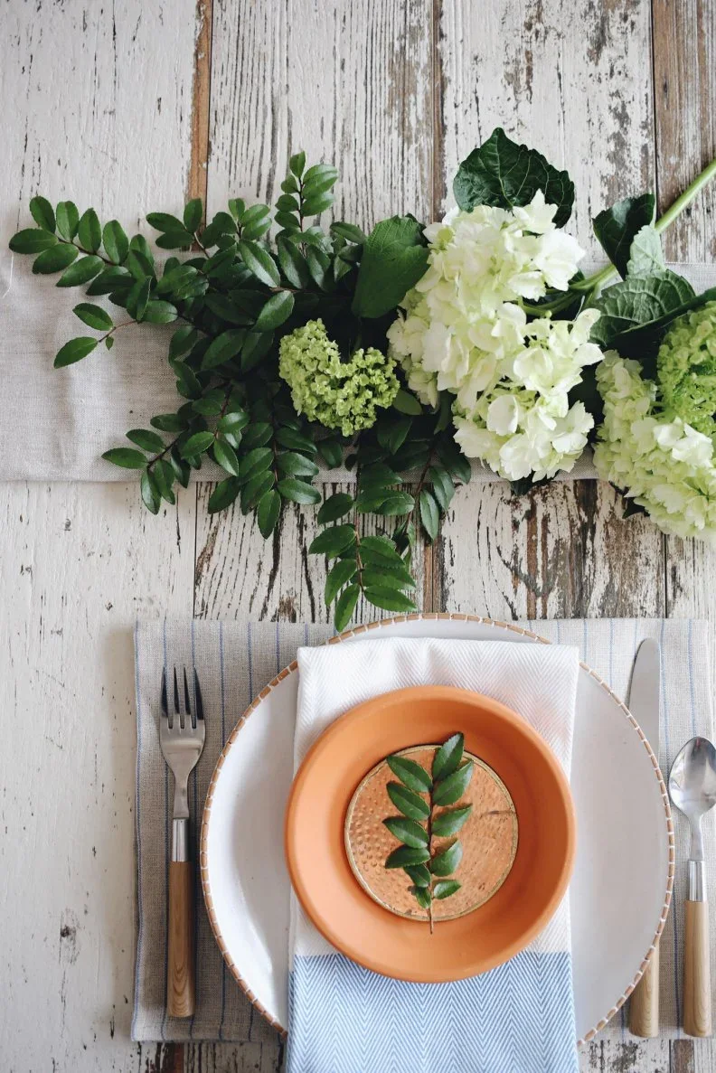 A green sprig lies on a small textured orange plate above a white napkin beside green-and-white flowers on pale wooden boards