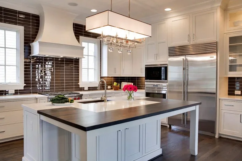 White framed cabinets and a pale island with darker seating edge sit beside dark horizontal backsplash tiles and a shaped white hood under a rectangular hanging light
