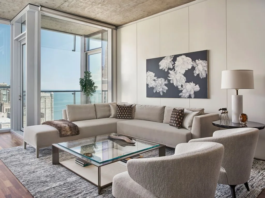 Living room with pale wall panels, a concrete ceiling, a taupe sectional, rounded armchairs and a glass-topped coffee table.