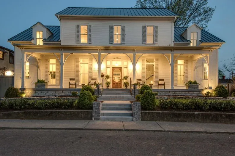 White farmhouse at dusk with illuminated front porch, central door, planted beds and stair approach