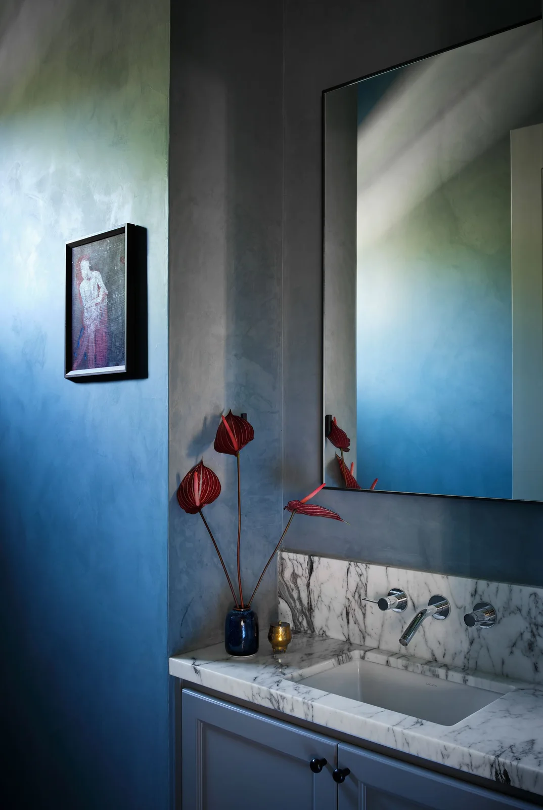 A large plain rectangular mirror hangs over a pale veined counter and blue cabinet fronts against blue-gradient walls beside a small vase of red flowers