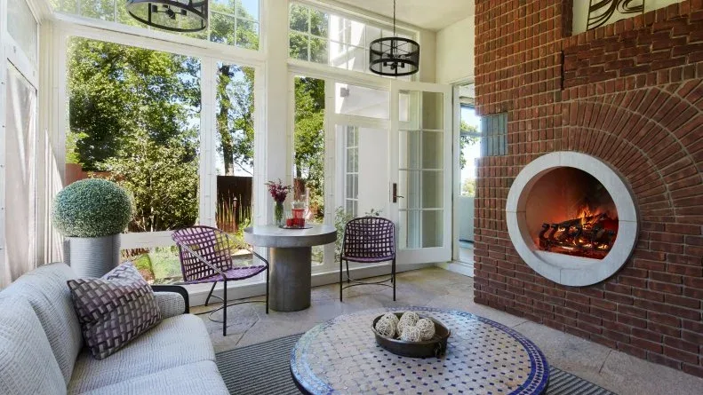 Red brick fireplace wall with round pale-edged opening beside large windows and gray sofa
