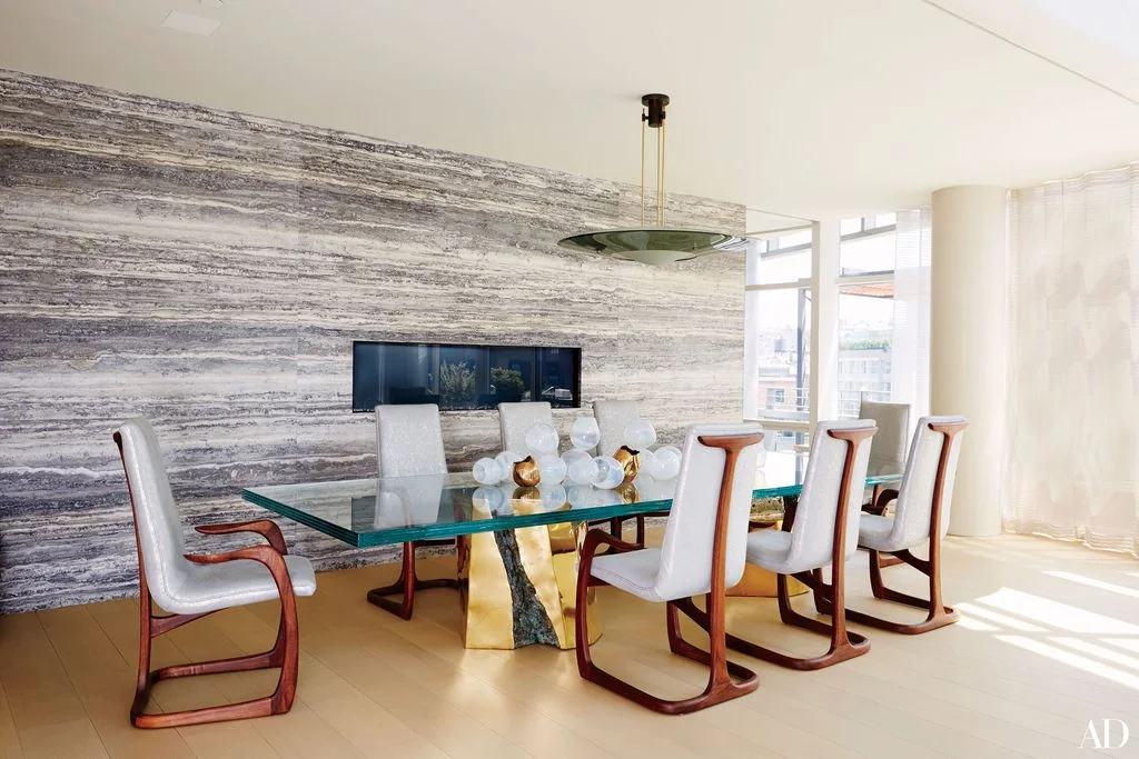 A narrow fireplace opening sits within a horizontally striped gray wall behind a transparent dining table and pale wooden-framed chairs