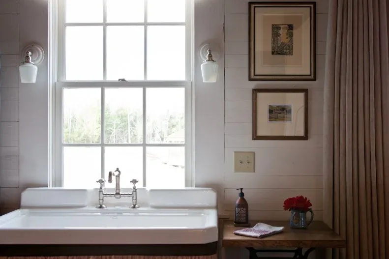 White farmhouse powder room with broad wall-mounted sink under window, framed pictures and small wood table