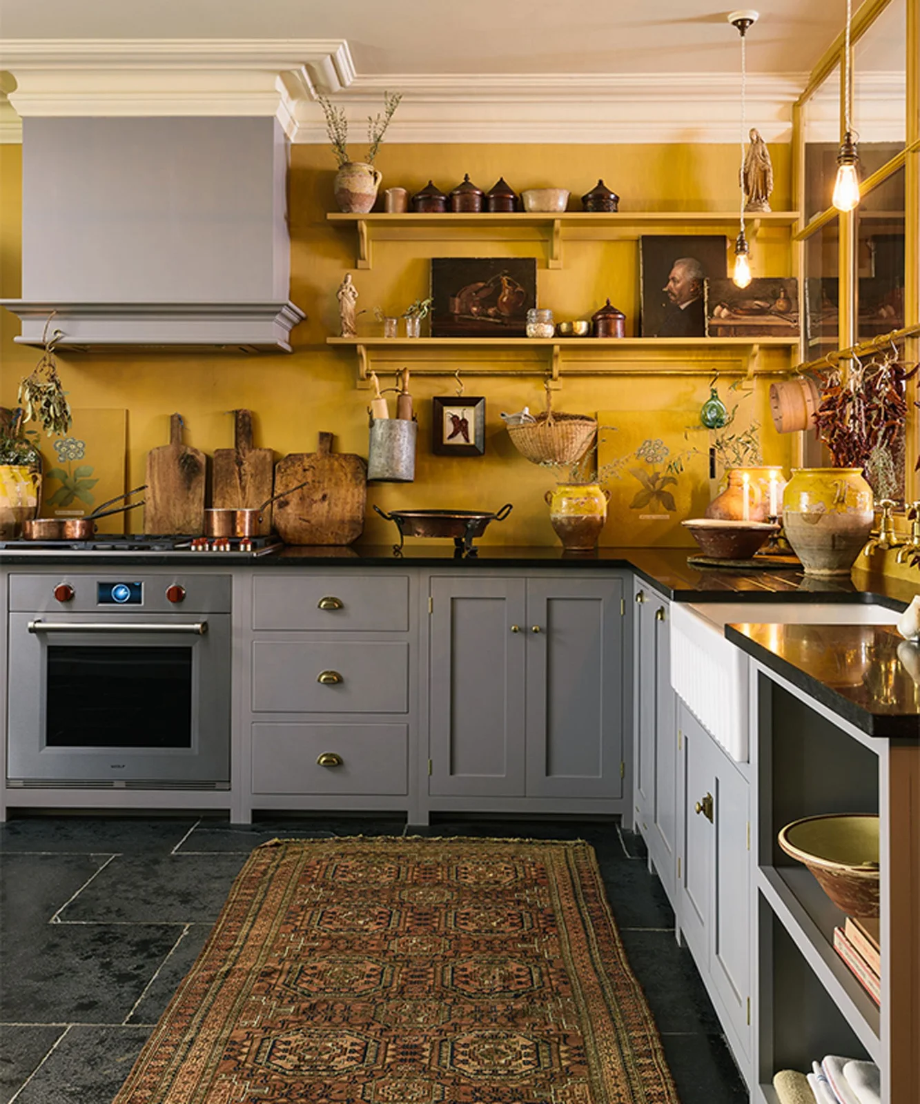 Pale gray paneled cabinets and drawers sit beneath yellow walls, shelves and hanging cookware beside a white apron-front sink
