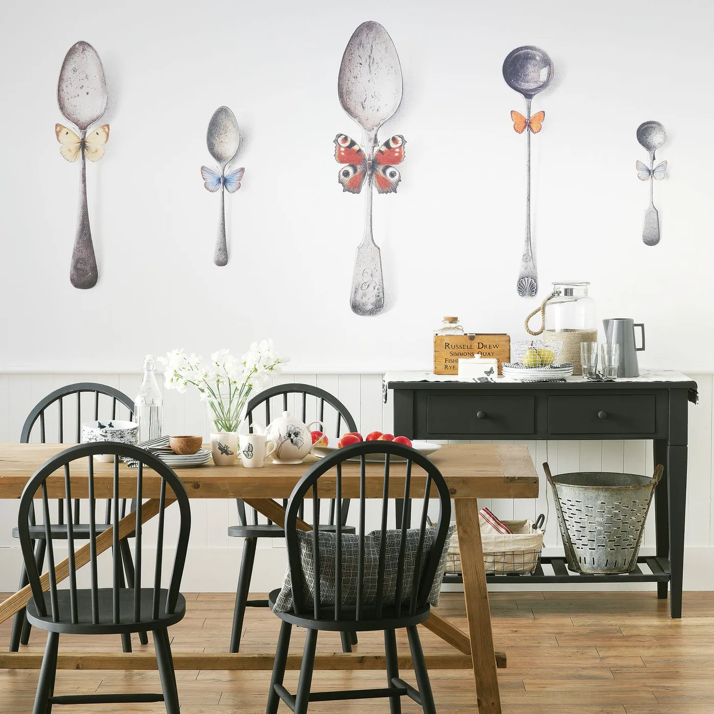 Cottage dining area with black spindle-back chairs, wooden table and bench, a dark side table and a large spoon-and-butterfly wall mural.