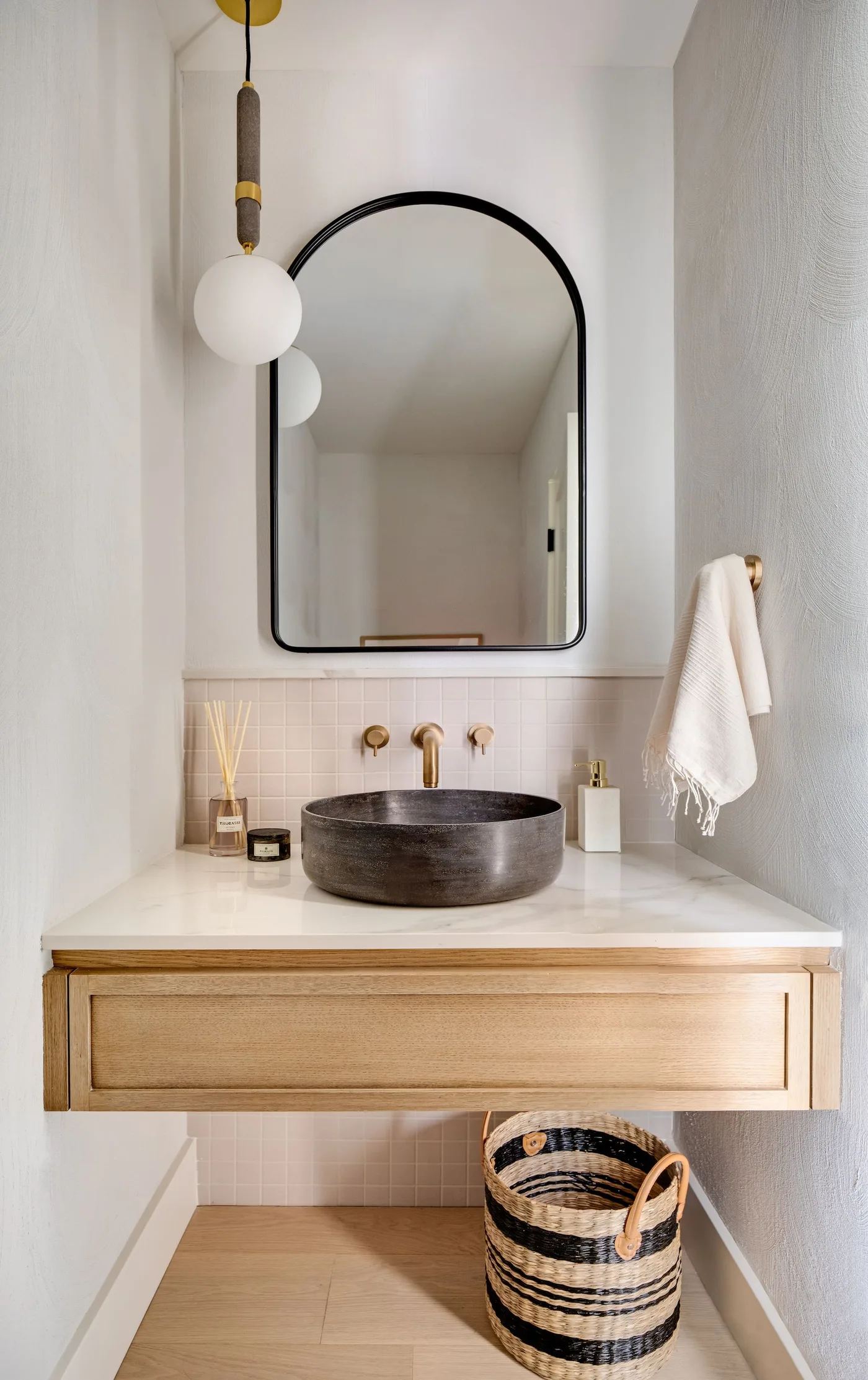 A floating pale wood drawer vanity holds a dark round basin beneath a black arched mirror beside a globe pendant, pale wall tiles and a woven basket