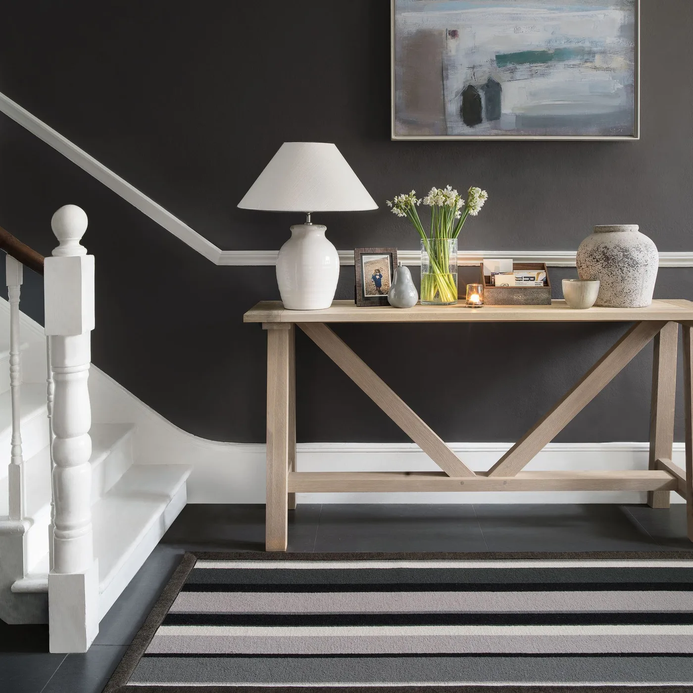 Broad black, gray and pale striped runner beneath a wood cross-braced console against a dark stair wall