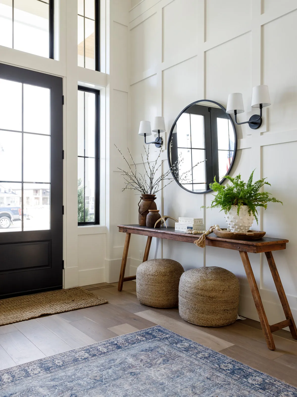 A wooden console holds plants and vessels beneath a round mirror beside two woven poufs, white paneled walls and a black glazed front door