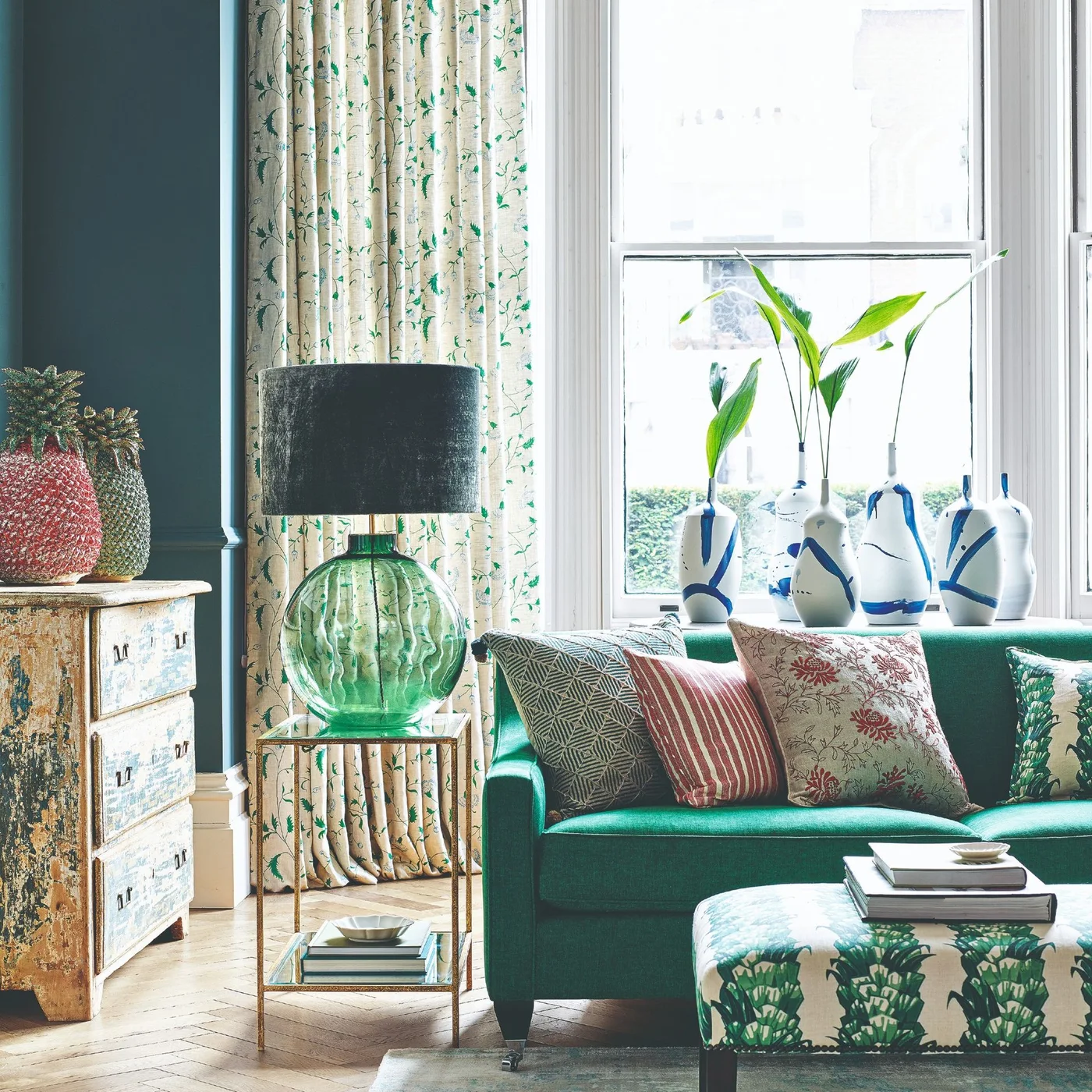 A compact emerald green sofa sits beside a green glass lamp, leafy curtains and a patterned footstool