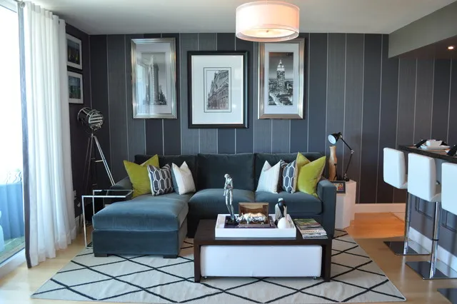 A blue sectional with pale and lime cushions sits against dark paneling above a pale geometric rug and square upholstered ottoman.