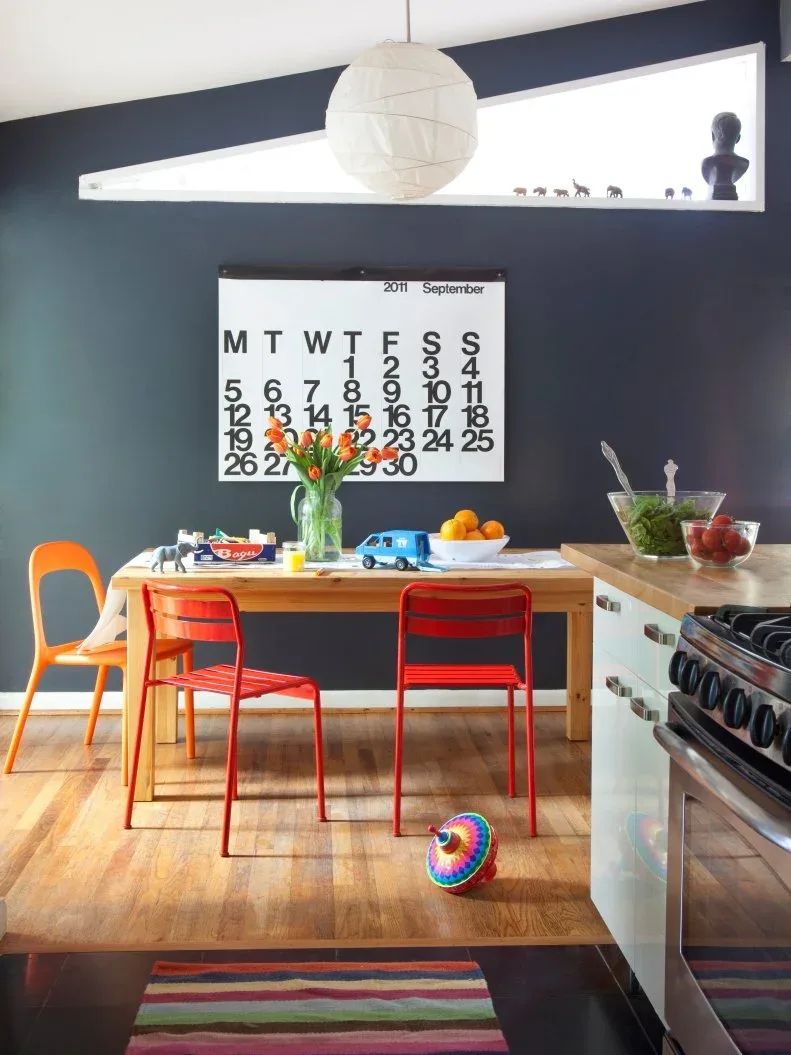 Dining nook with a dark blue-gray wall, triangular window, large calendar, wooden table, orange and red chairs, and a white paper pendant.