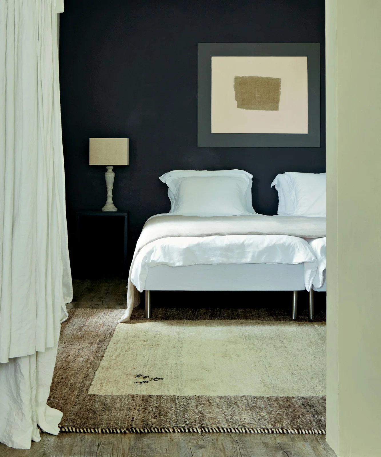 White bedding sits against a dark headboard wall beside pale curtains, a small bedside lamp and layered light floor coverings