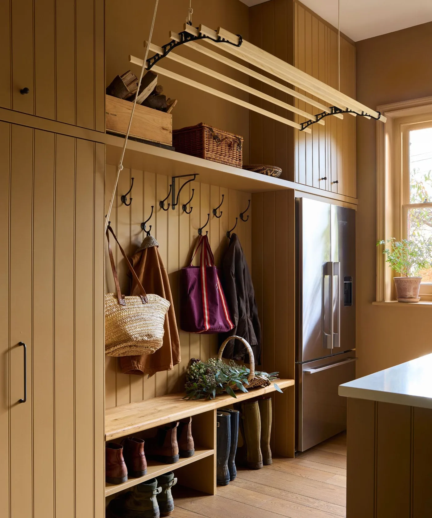 Warm brown paneled boot room with ceiling-suspended slatted rack, coat hooks, wood bench and open boot cubbies