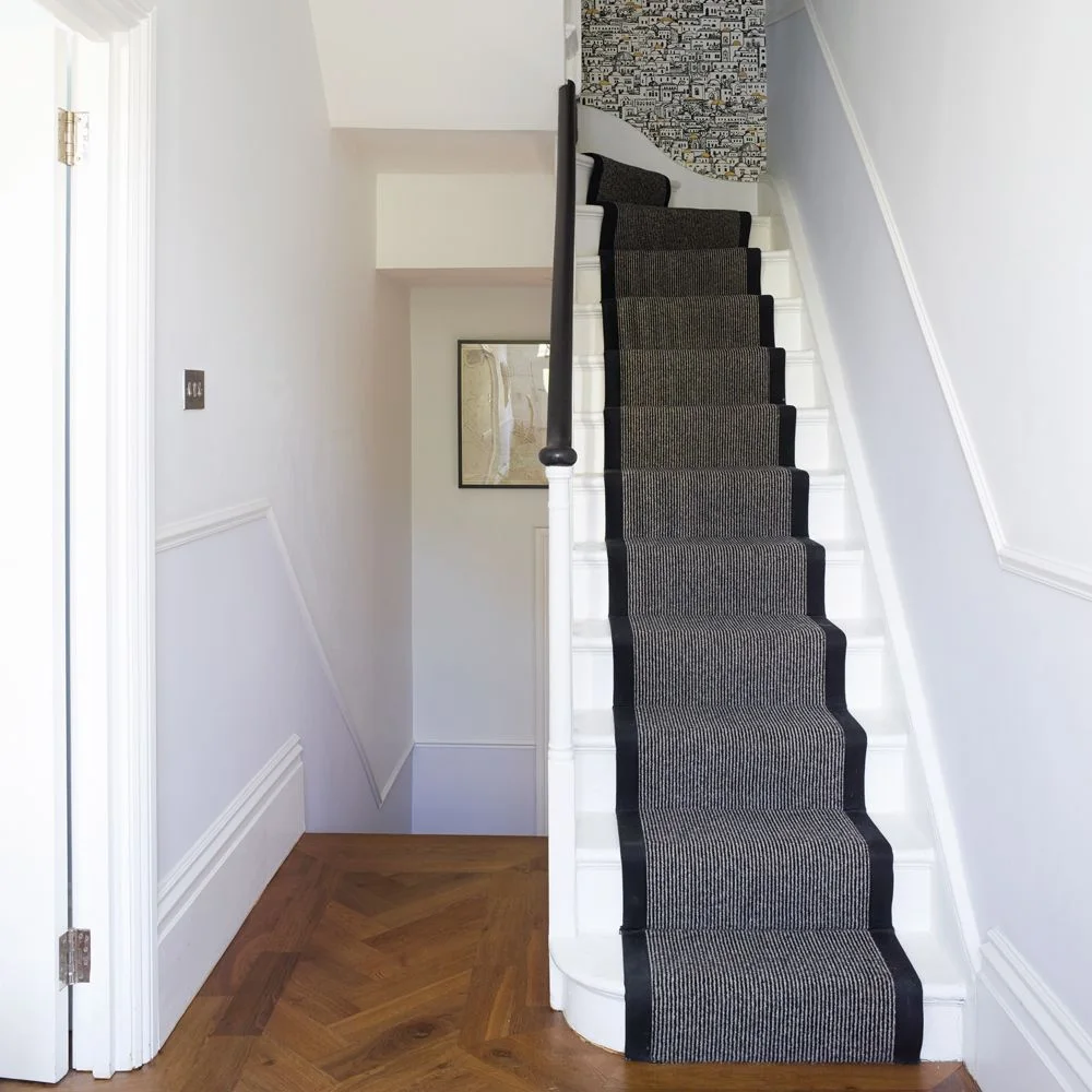 A narrow white hallway has a dark gray stair runner with black borders below a black handrail and patterned wall at the upper landing