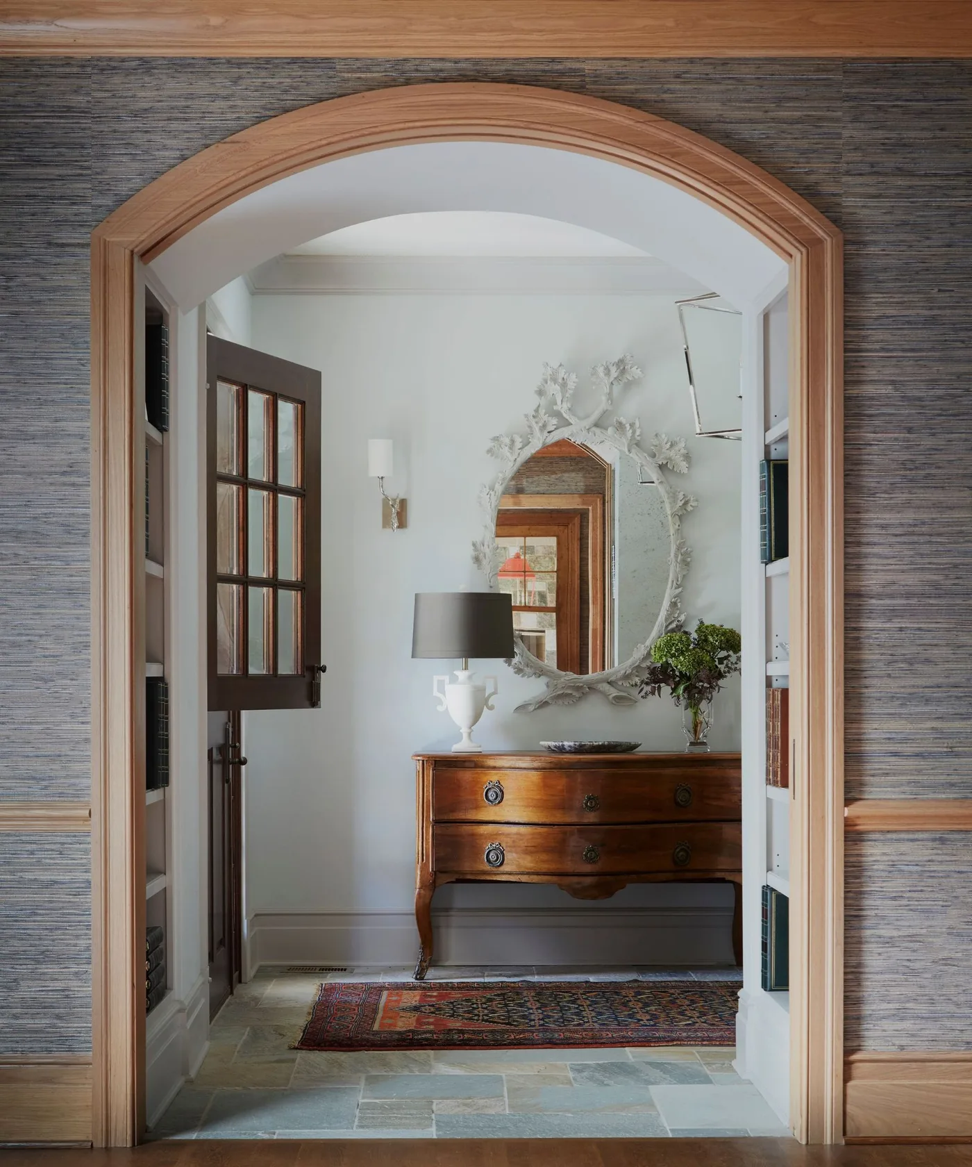 Entrance viewed through wooden arch with warm drawer console, pale wall, decorative oval mirror, gray lamp and patterned runner.