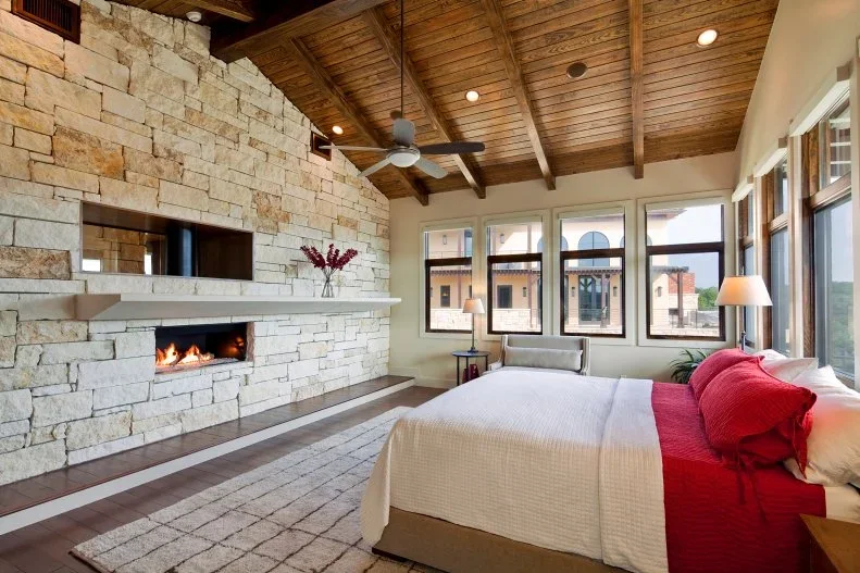 A broad pale textured fireplace wall with a long horizontal opening sits beneath an angled wood ceiling beside a bedroom with white bedding and red pillows