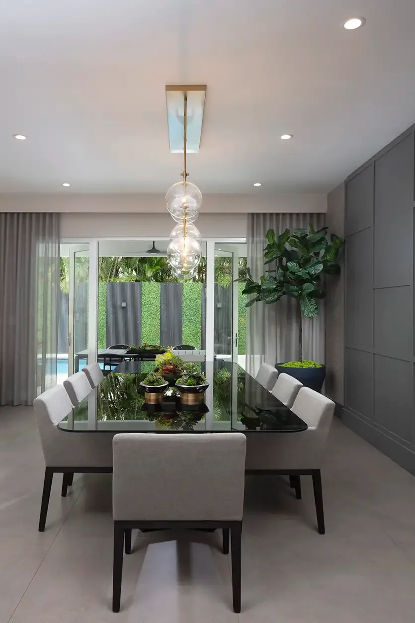 A dark glass dining table and pale upholstered chairs sit beneath clear globe pendants beside a gray paneled wall and light curtains