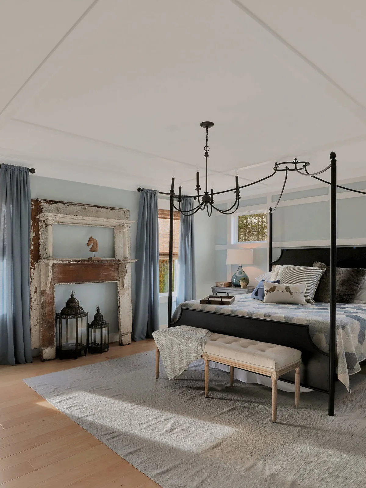 A black four-poster bed stands in a pale room with exposed ceiling details and a rustic mantel.