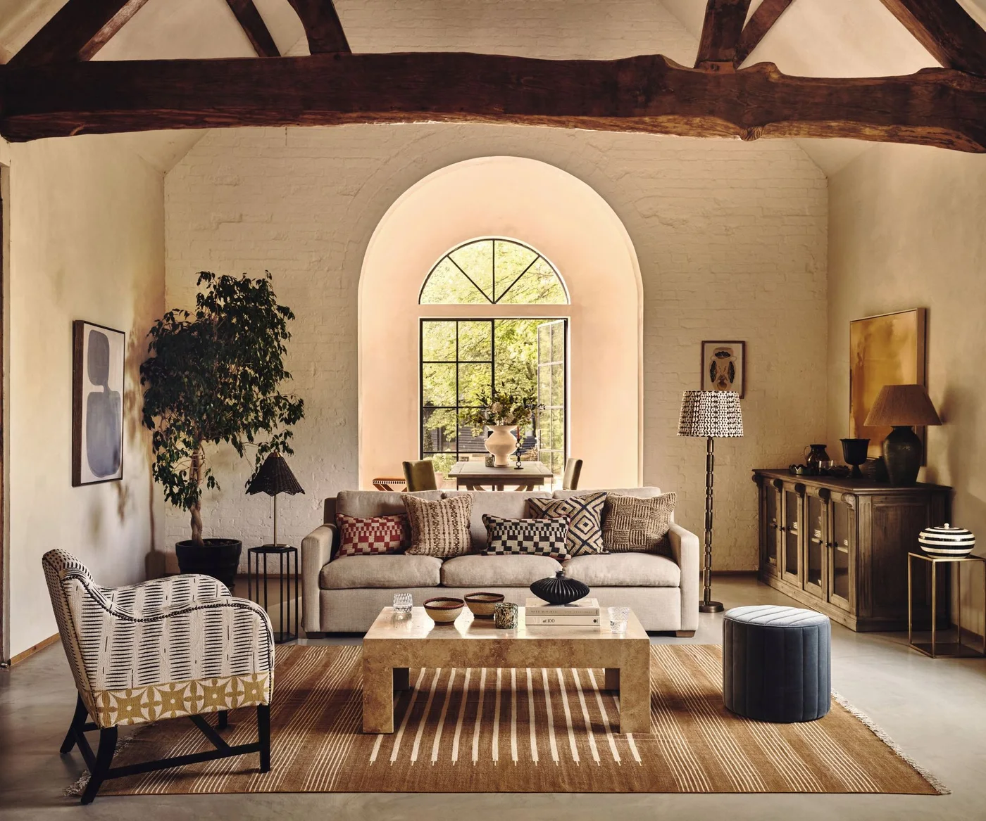 A deep arched living-room window rises behind a pale sofa and patterned chair beside dark beams, a wooden coffee table and warm striped rug