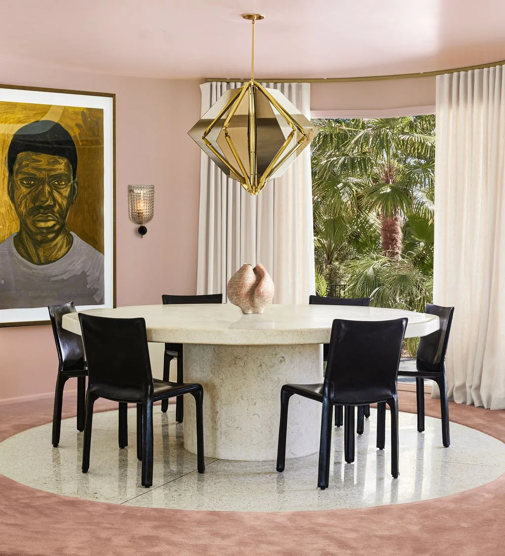A pale round pedestal dining table and black chairs sit beneath a geometric gold-toned pendant beside soft pink walls and white curtains