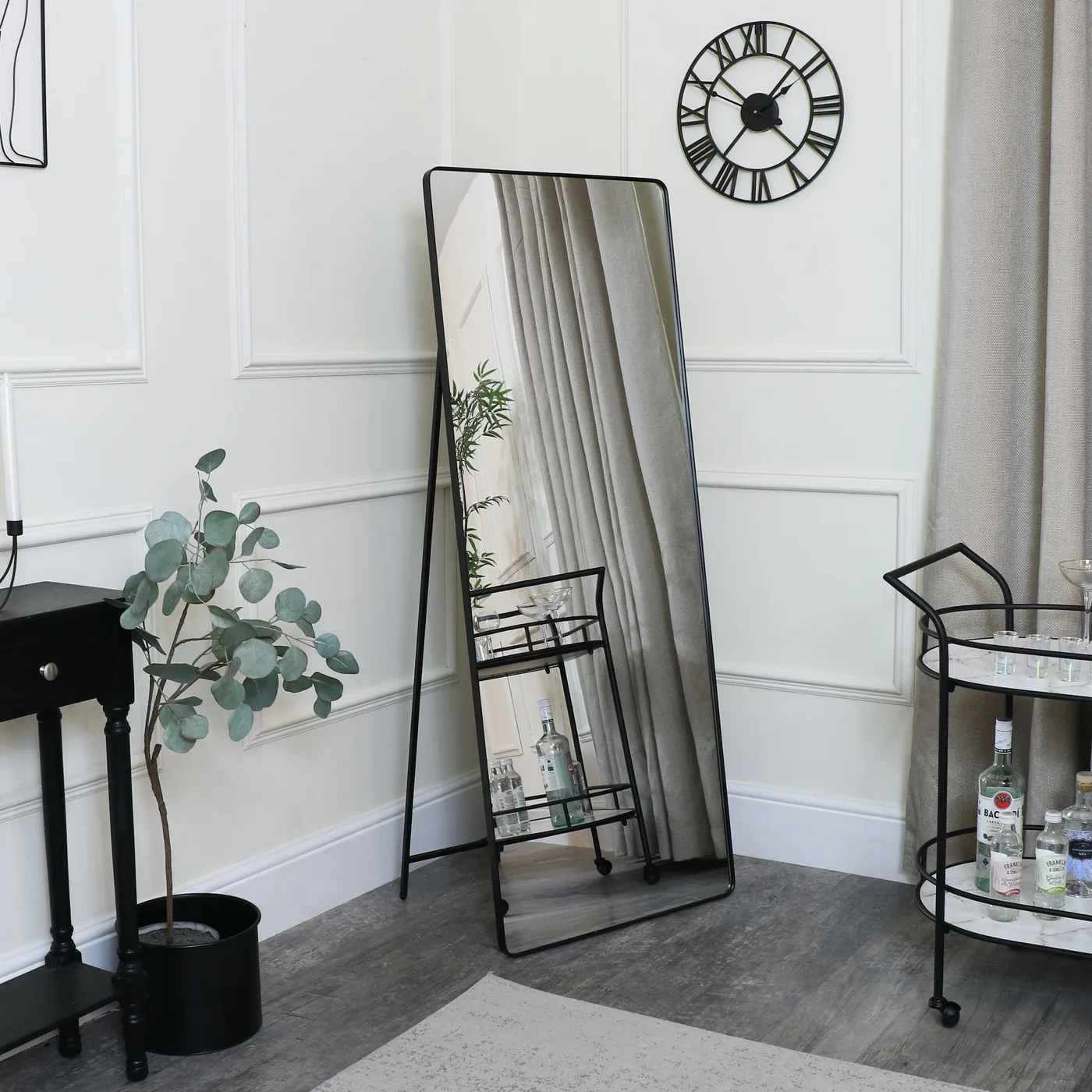 Tall narrow black-framed mirror beside a black drinks cart against a pale paneled wall