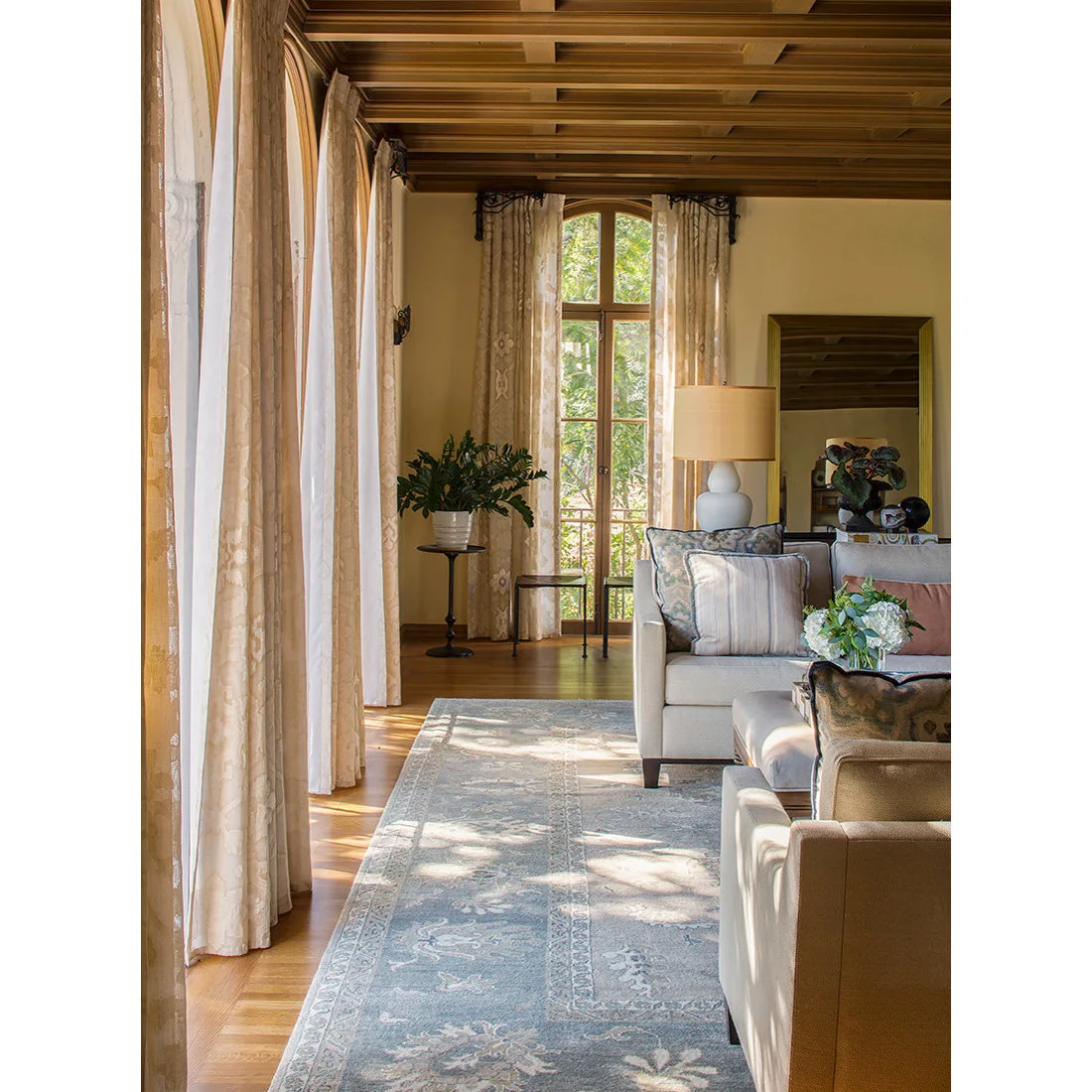 A wood-patterned ceiling sits above pale living-room seating, a fine patterned rug and long curtains beside tall glazed doors and a large mirror