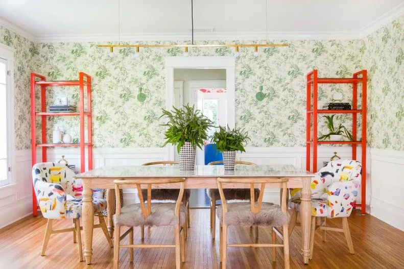 Wooden dining chairs with fluffy seat coverings beside multicolored upholstered end chairs and a wooden table, against green floral wallpaper and red shelves.