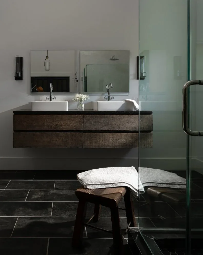 A dark-tiled bathroom floor meets a floating wood vanity with two white bowl sinks below a wide mirror beside a glass shower screen