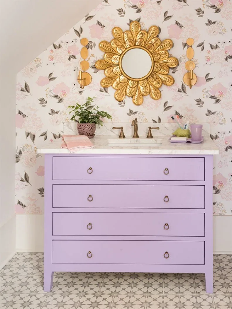 Powder room with pale floral wallpaper, lavender drawer vanity, gold petal-shaped mirror, golden wall lights, pale basin counter and patterned floor tiles.