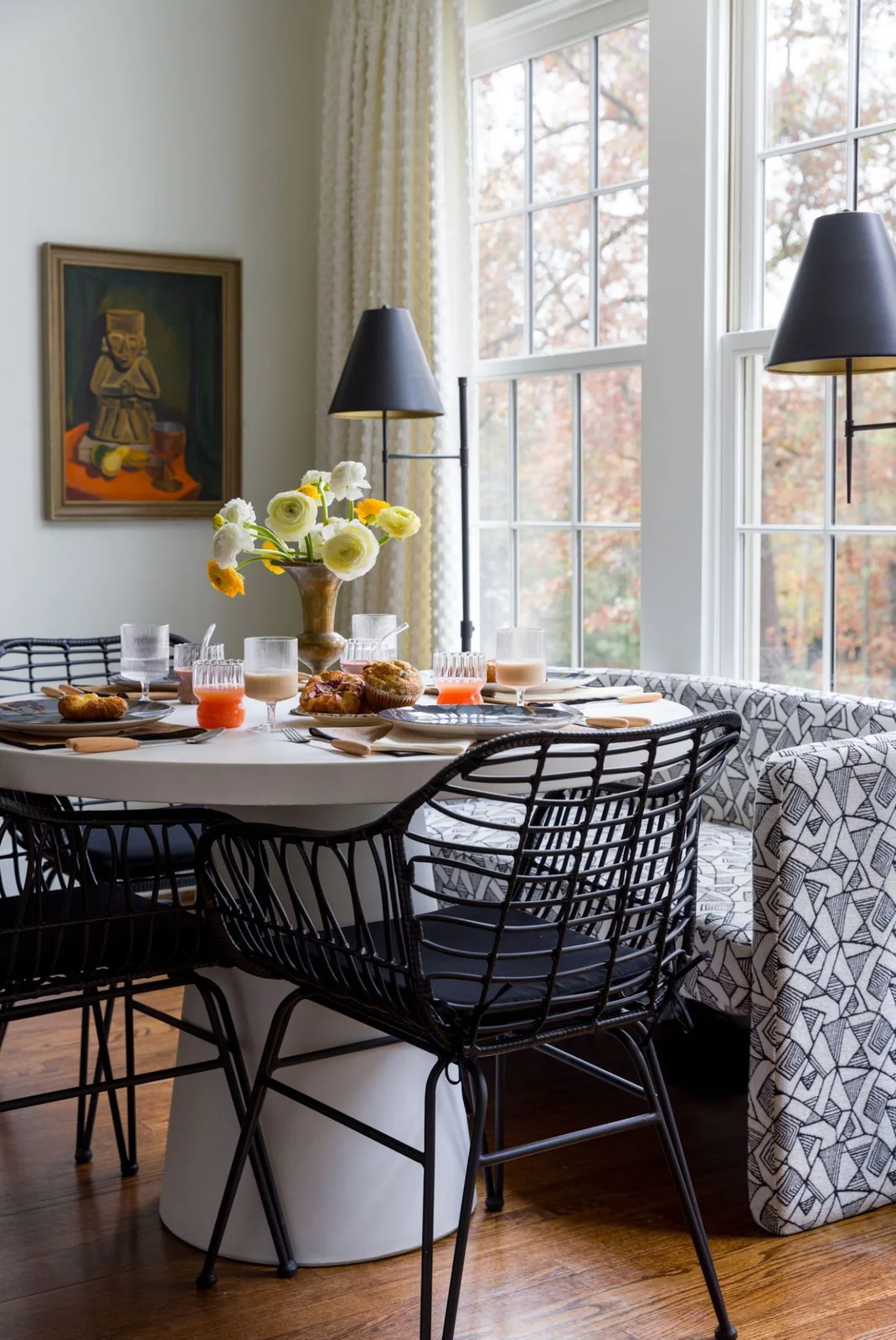 Black open dining chairs surround a white pedestal table beside a black-and-white patterned window bench and dark-shaded lights